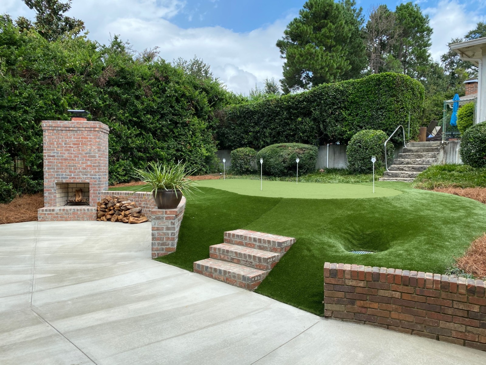 An outdoor brick fireplace and chimney with a stacked woodpile, brick steps and clipped hedges, opening onto a green putting surface