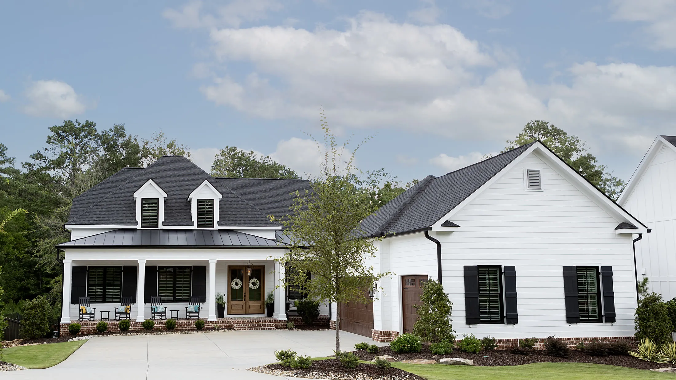 White farmhouse with a standing-seam porch roof and twin dormers, fronted by a concrete drive, a young maple and freshly mulched foundation beds